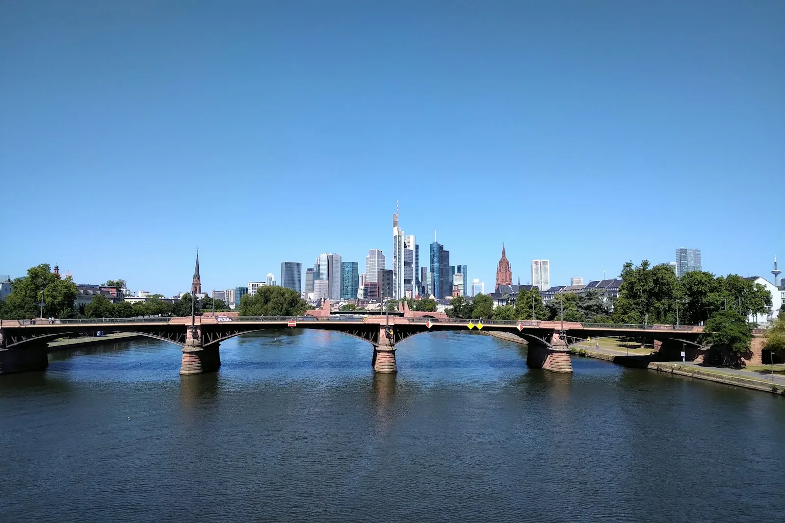 Blick über den Main auf die Ignatz-Bubis-Brücke, dahinter die Hochhäuser der Skyline unter blauem Himmel