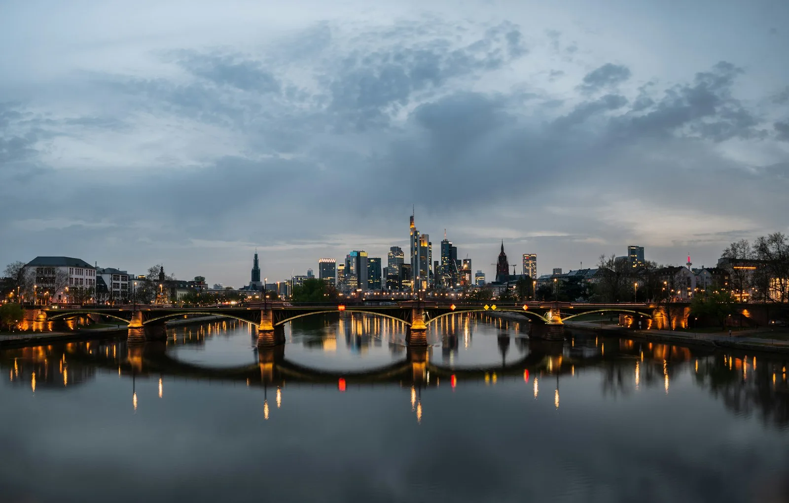 Die beleuchtete Frankfurter Skyline in der Abenddämmerung, im Vordergrund eine Mainbrücke mit Lichterreihen